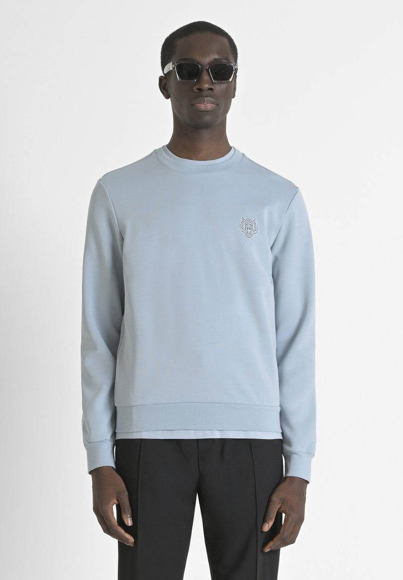 Lichtblauwe sweatshirt van zacht materiaal, met een ronde hals en een geborduurd logo. Combinatie met zwarte broek en zonnebril.