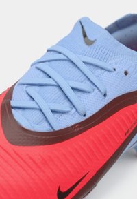 Les baskets Nike présentent une tige en tricot bleu clair avec des lacets fins, une semelle texturée rouge vif, et un accent marron foncé autour de la zone des orteils.