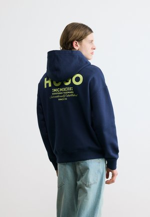 Jeune personne portant un sweat à capuche Hugo bleu marine avec un texte vert citron dans le dos et un jean bleu clair, se tenant devant un fond blanc, regardant sur le côté.