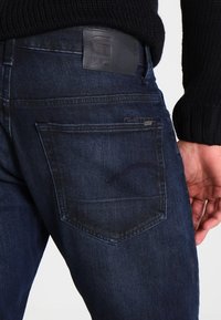 Tmavě modré denimové džíny mají koženou nášivku na pasu s logem. Zadní kapsa má decentní šev.