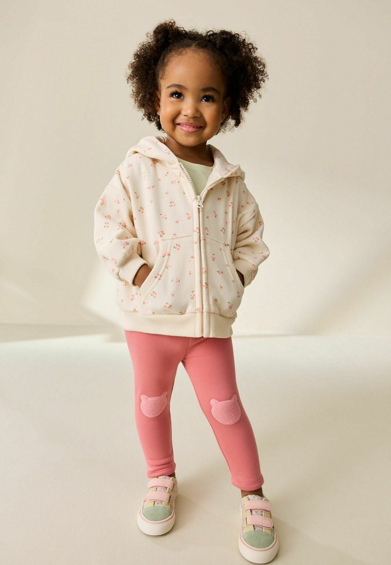 Jasnobeżowa bluza z kapturem na zamek błyskawiczny z kwiatowym wzorem, zestawiona z różowymi legginsami z łatkami na kolanach w kształcie misiów oraz wielokolorowymi trampkami.