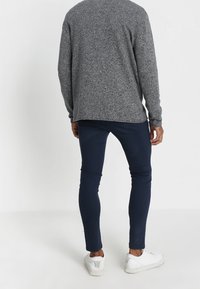 Harmaa, teksturoitu neulepaita, jossa on pitkät hihat, on päällä tummansiniset slim fit -housut ja valkoiset lenkkarit. Tausta on tasaisen valkoinen.