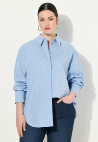 Camicia azzurro chiaro con colletto, maniche lunghe e vestibilità rilassata, realizzata in tessuto liscio. Indossata con pantaloni in denim scuro.