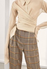 Chemisier beige transparent à col et boutons, associé à un pantalon large à carreaux aux tons noir, blanc et orange.