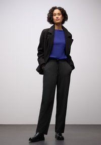 Zwarte jas over een blauwe trui, gecombineerd met grijze broek en zwarte enkellaarzen. Soepele texturen en een op maat gemaakte, ontspannen pasvorm.