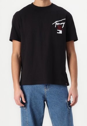 Camiseta de algodón negra de manga corta. Presenta un logotipo en blanco en el pecho y acentos rojos. Combinada con unos vaqueros de denim azul claro.