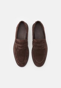 Brune semskede loafers med en bred rem over vampen, rund tå og sydd detaljer langs kantene. Myk tekstur og slank design.