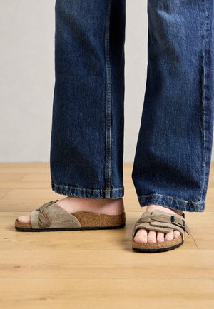 Voeten in beige suède Birkenstock sandalen met kurken zolen en brede banden, gecombineerd met donkerblauwe denim jeans op een houten vloer.