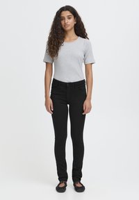 Joven con cabello rizado, vistiendo una blusa de manga corta gris claro, jeans ajustados negros y zapatos de ballet negros, de pie sobre un fondo blanco.