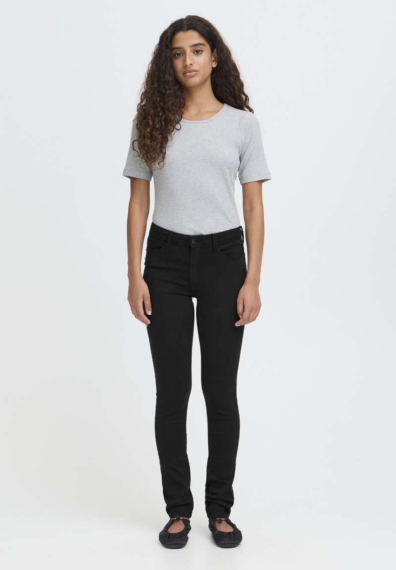 Joven con cabello rizado, vistiendo una blusa de manga corta gris claro, jeans ajustados negros y zapatos de ballet negros, de pie sobre un fondo blanco.