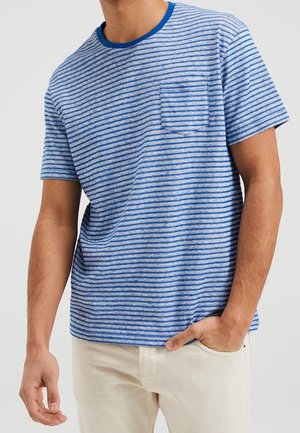 Homme portant un t-shirt à manches courtes rayé bleu et blanc avec une poche poitrine et un pantalon beige clair, debout avec une main dans la poche.