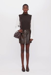 Top in maglia senza maniche marrone, camicia bianca a maniche lunghe sotto, gonna nera in pelle con zip, borsa marrone, ballerine nere con dettagli decorativi.