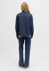 Jeans jakna s klasičnim krojem, temno modre barve, z gumbi ter dvema žepoma na prsih. V kombinaciji z širokimi temnimi kavbojkami.