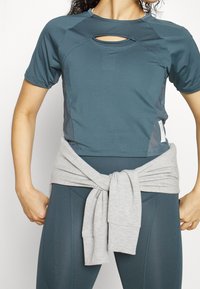 Kvinna klädd i turkos atletisk crop top och leggings med en ljusgrå sweatshirt knuten runt midjan, stående framför en vit bakgrund.