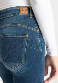 Jeans in denim blu con una etichetta in pelle con la scritta "LC", cuciture gialle e un motivo ricamato tonale sulla tasca posteriore.