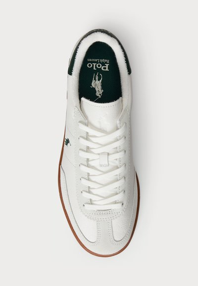 Baltas sneakers ar teksturētu ādu, gumijas zoli un zaļu iekšzoli ar logotipu. Šņores ir baltas, un tajos ir dekoratīvas šūšanas detaļas.