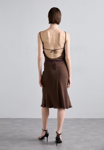 Vestito slip in satin marrone con design a schiena bassa e spalline regolabili; presenta un orlo svasato e décolleté neri con cinturini.