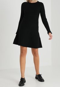 Abito di maglia nera con maniche lunghe, scollatura rotonda e orlo svasato. Tessuto testurizzato e design minimalista. Indossato con scarpe nere.
