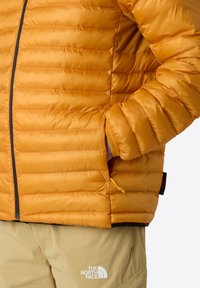 Izolovaná žlutá péřová bunda s horizontálním prošíváním, se zipovým zapínáním a bočními kapsami. V kombinaci s světle béžovými kalhotami s logem.