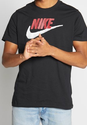 Osoba ubrana w czarną koszulkę Nike z czerwonym logo i białym znakiem, z rękami splecionymi razem, w jasnoniebieskich dżinsach.