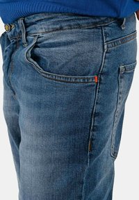 Jeans in denim blu con design a gamba dritta, cuciture visibili e accenti arancioni sulla tasca. La texture appare robusta e durevole.