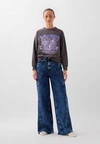 Felpa grigia con stampa grafica, abbinata a jeans in denim blu a vita alta e gamba larga. Scarpe casual completano l'outfit.