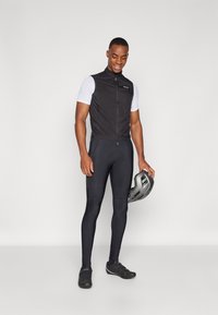 Gilet aerodinamico nero con zip frontale, abbinato a leggings ciclistici aderenti neri. Il modello tiene un casco da ciclismo grigio.