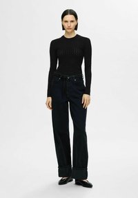 Zwarte langslijf top met een gestructureerd patroon, gecombineerd met donkere high-waisted flared jeans met een opgerolde zoom en een strik ceintuur detail.