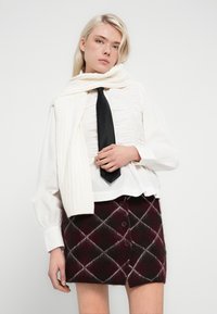 Γυναίκα που φοράει λευκό πουκάμισο με ρυτιδώσεις, μαύρη γραβάτα, λευκό πουλόβερ περασμένο στους ώμους και κουμπωτή φούστα σε βυσσινί καρό.