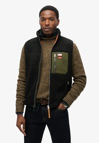 Sort fleece vest med en grøn lynlåslomme, orange accent lynlås og et patch. Båret over en brun tekstureret sweater og en blå ternet skjorte.