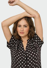 Blouse noire à pois blancs, à manches courtes et avec un design à boutons sur le devant. Tissu doux avec une coupe décontractée et un col rond.