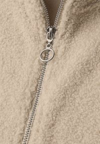 Beżowa kurtka z polaru o miękkiej, fakturowanej powierzchni oraz metalowym zamku błyskawicznym z okrągłym uchwytem. Zbliżenie na otwarcie zamka.