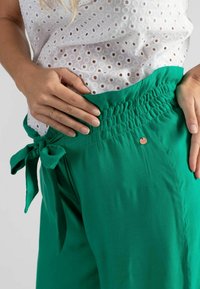 Groene, loszittende broek met een smocktail en strikdetail, en een kleine metalen accent. Zacht materiaal met een gladde textuur.