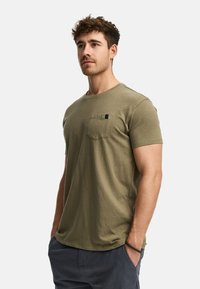 Camiseta de algodón verde oliva con mangas cortas, cuello redondo y un bolsillo en el pecho. Detalles de costura notables y ajuste casual.