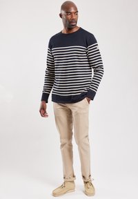 Marineblå og hvide stribede langærmede sweater, crew neck design, parret med beige bukser og lyse beige sko, sat mod en ensfarvet baggrund.