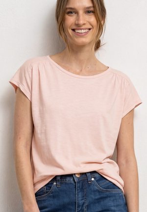 Femme souriante portant un t-shirt rose clair à manches courtes rentré dans un jean bleu taille haute, debout devant un fond blanc.