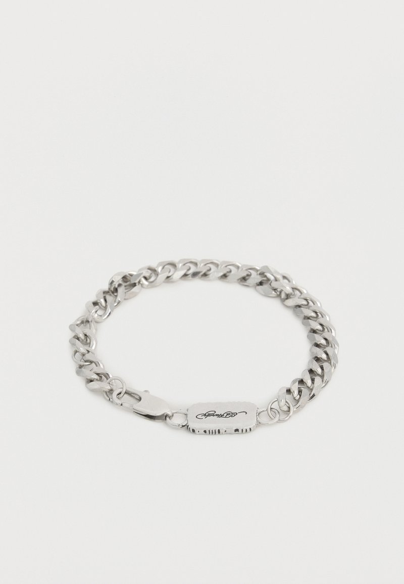 Sølvfarvet kædearmbånd med en lås i hummerklo-stil og et rektangulært mærke med en indgraveret, stiliseret signatur.