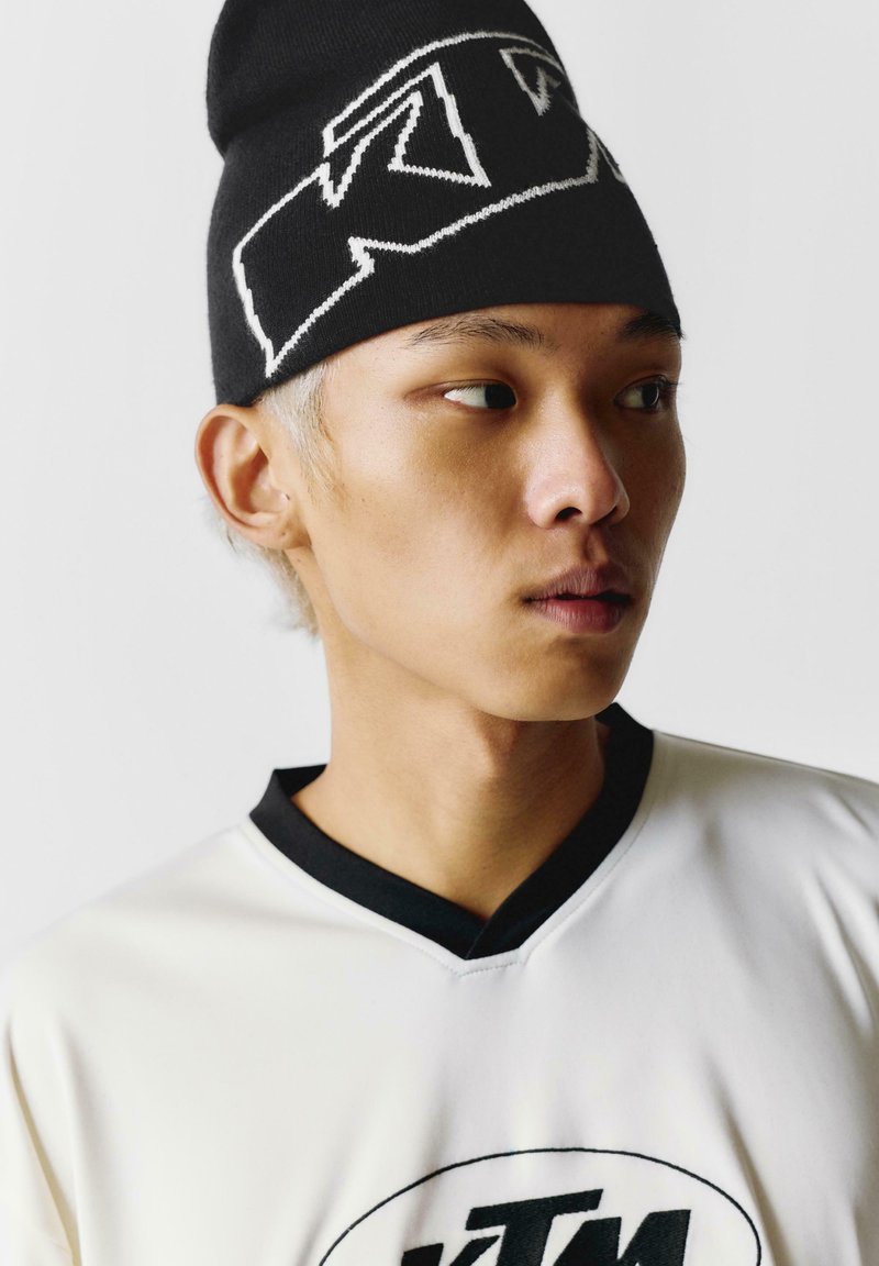 Black knit beanie with white logo, worn with a white shirt featuring a black collar and a circular logo at the center. Close-up portrait.