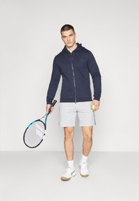 Felpa zip con cappuccio blu navy, abbinata a pantaloni corti grigi. Tiene una racchetta da tennis e una palla, indossando sneakers bianche e calzini alla caviglia. Design minimalista.