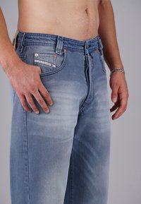 Close-up of a person wearing light blue denim jeans with faded details, showing their bare midriff and hands resting by their sides.