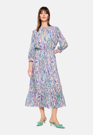 Frau trägt ein langärmliges Midikleid mit pastellfarbenem abstraktem Muster, tailliertem Schnitt, Rüschen am Saum und grünen, niedrighackigen Slingback-Schuhen.