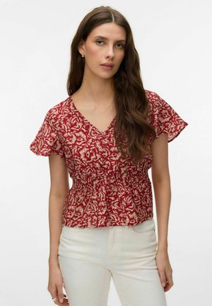 Femme aux longs cheveux bruns portant un chemisier rouge à fleurs avec un col en V et des manches courtes à volants, ainsi qu'un pantalon blanc, debout devant un fond blanc.