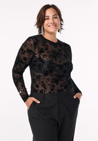 Sort transparent langærmet top med blomster-velourmønster, parret med tætsiddende sorte bukser. Modellen har kort hår og øreringe.
