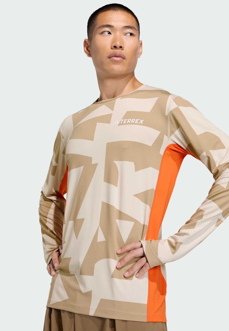 Maglietta sportiva a maniche lunghe beige con motivi astratti, dotata di un pannello laterale arancione e logo "TERREX" sul petto. Texture liscia.