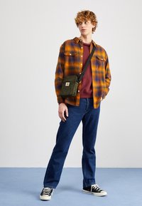 Nuori mies, joka on pukeutunut oranssi-purppuraan ruudulliseen paitaan, burgundinväriseen huppariin, sinisiin farkkuihin, mustiin lenkkikenkiin ja vihreään olkalaukkuun, seisoo sinisellä lattialla.