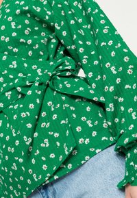 Blusa floral verde com padrões de flores brancas e castanhas, apresenta uma cintura amarrada, tecido texturizado e mangas longas com punhos franzidos.