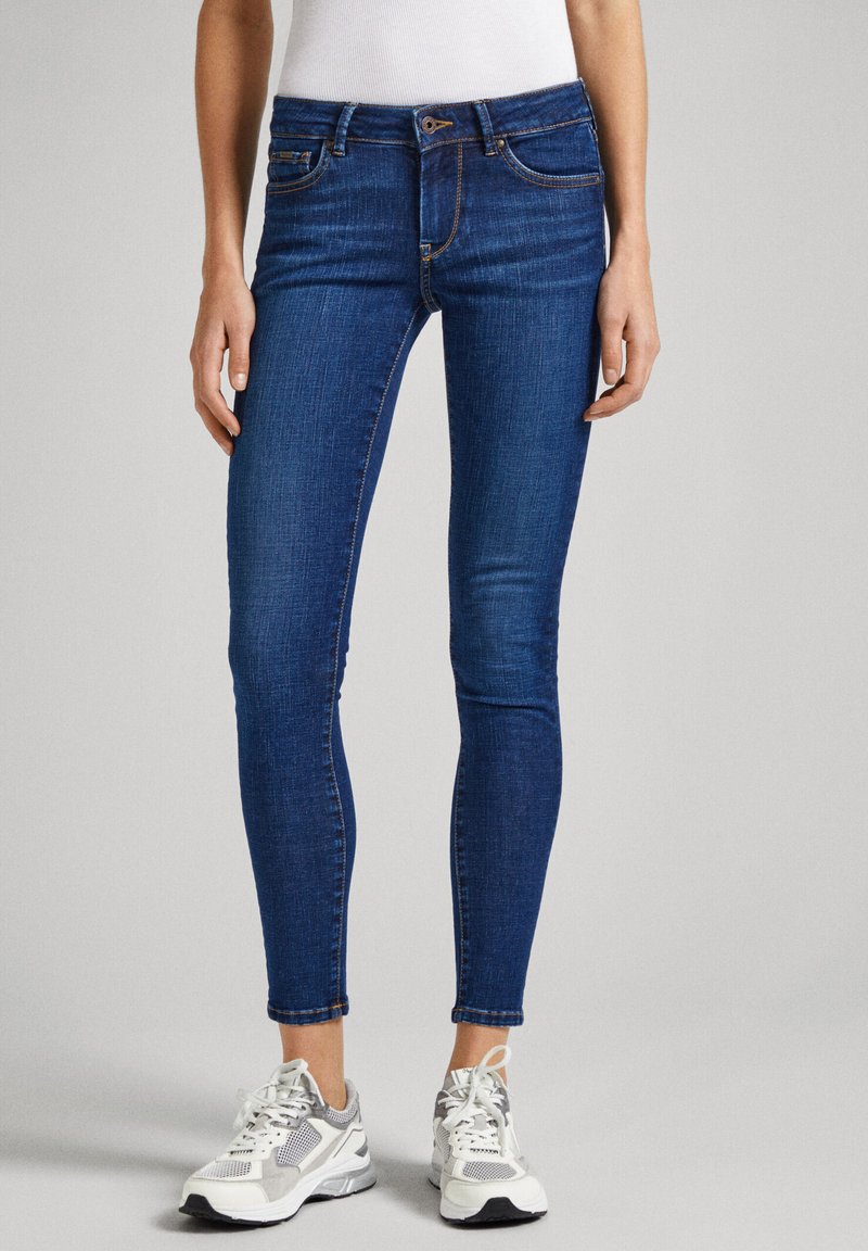 Jeans ajustados de mezclilla elástica en azul oscuro, con una cintura de altura media, cinco bolsillos y detalles de costura visibles. Combinados con zapatillas gris y blancas.