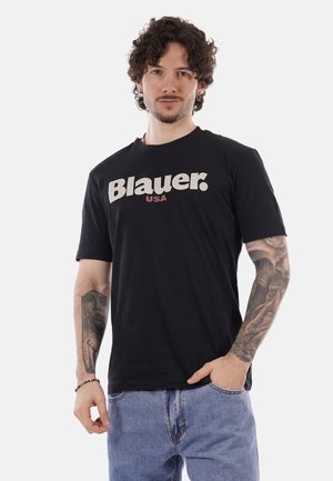 Uomo con capelli ricci, barba e tatuaggi, indossa una maglietta nera "Blauer USA" e jeans blu, in piedi con una mano in tasca.