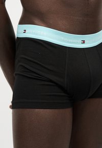 Κοντινό πλάνο ενός ατόμου που φοράει μαύρα μποξέρ με ανοιχτό μπλε μέση με τη στάμπα «Tommy Hilfiger».