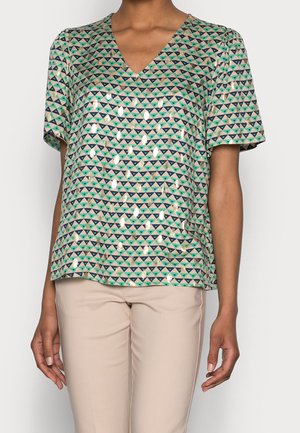 Vrouw draagt een kort mouwtjes V-hals top met groene en blauwe driehoekpatronen en glanzende gouden accenten, gecombineerd met beige broek.